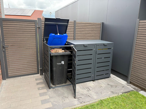 Kundenfoto: graue Alubert Mülltonnenbox neben Zaun mit geöffnetem Deckel und blauer 240-Liter-Tonne