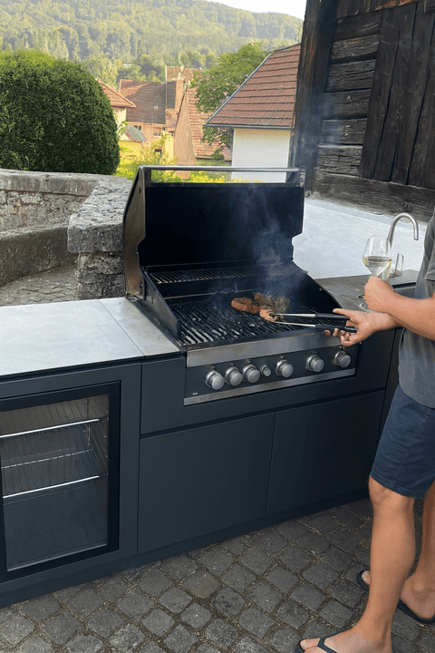 Person grilling in an outdoor kitchen with a view of a rustic landscape.